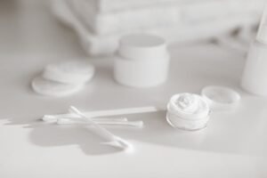 An open white moisturizer jar beside cotton swabs on a pale surface