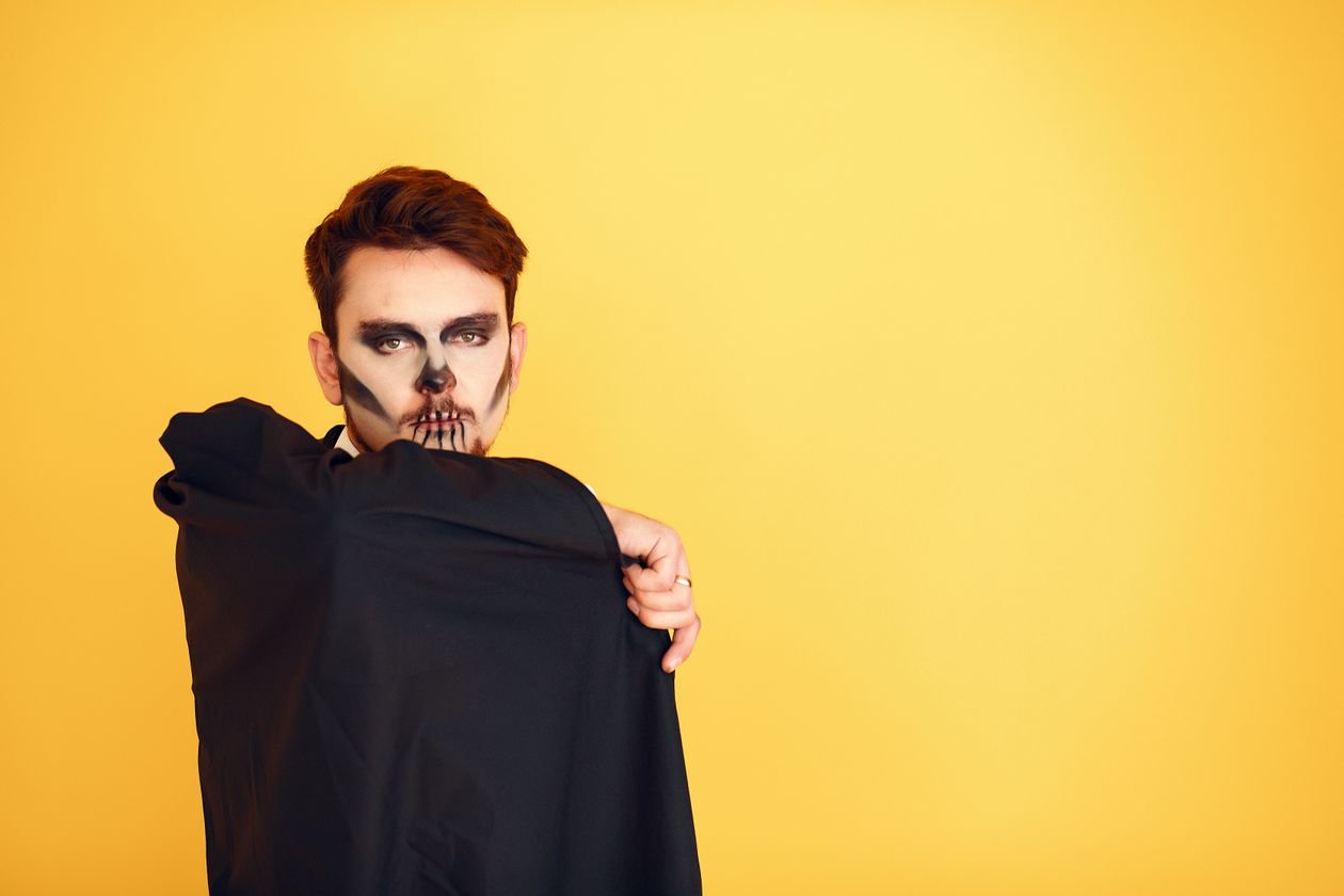 Man in black and white skull face paint and a black cape against a yellow background