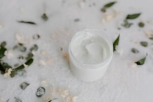 An open jar of moisturizer on a pale surface with leaves and petals around it